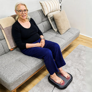Kvinne sitter i sofaen med begge føttene på fotmassasjemaskinen på gulvet