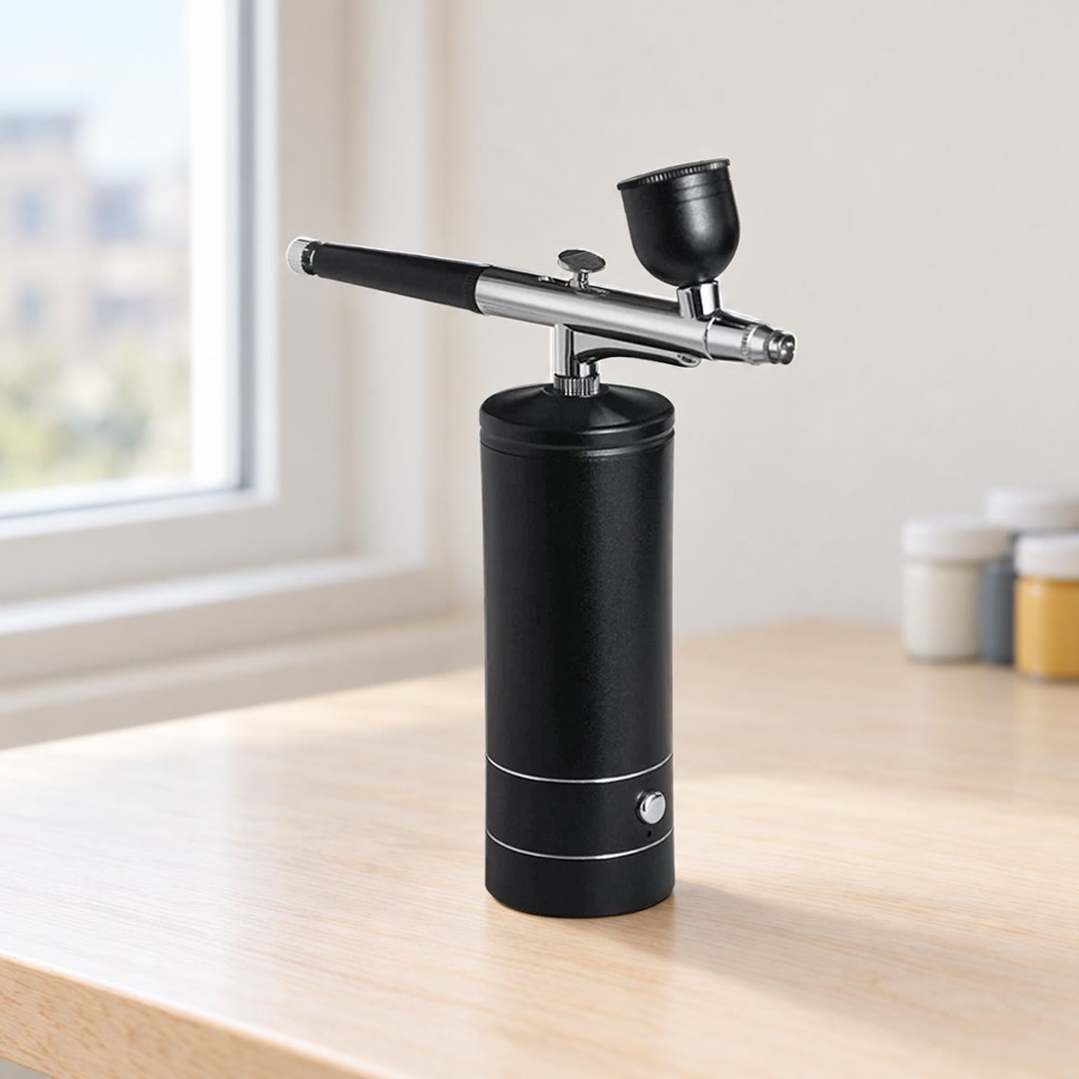 Svart airbrush med minikompressor på et lyst skrivebord i bjørk ved et vindu