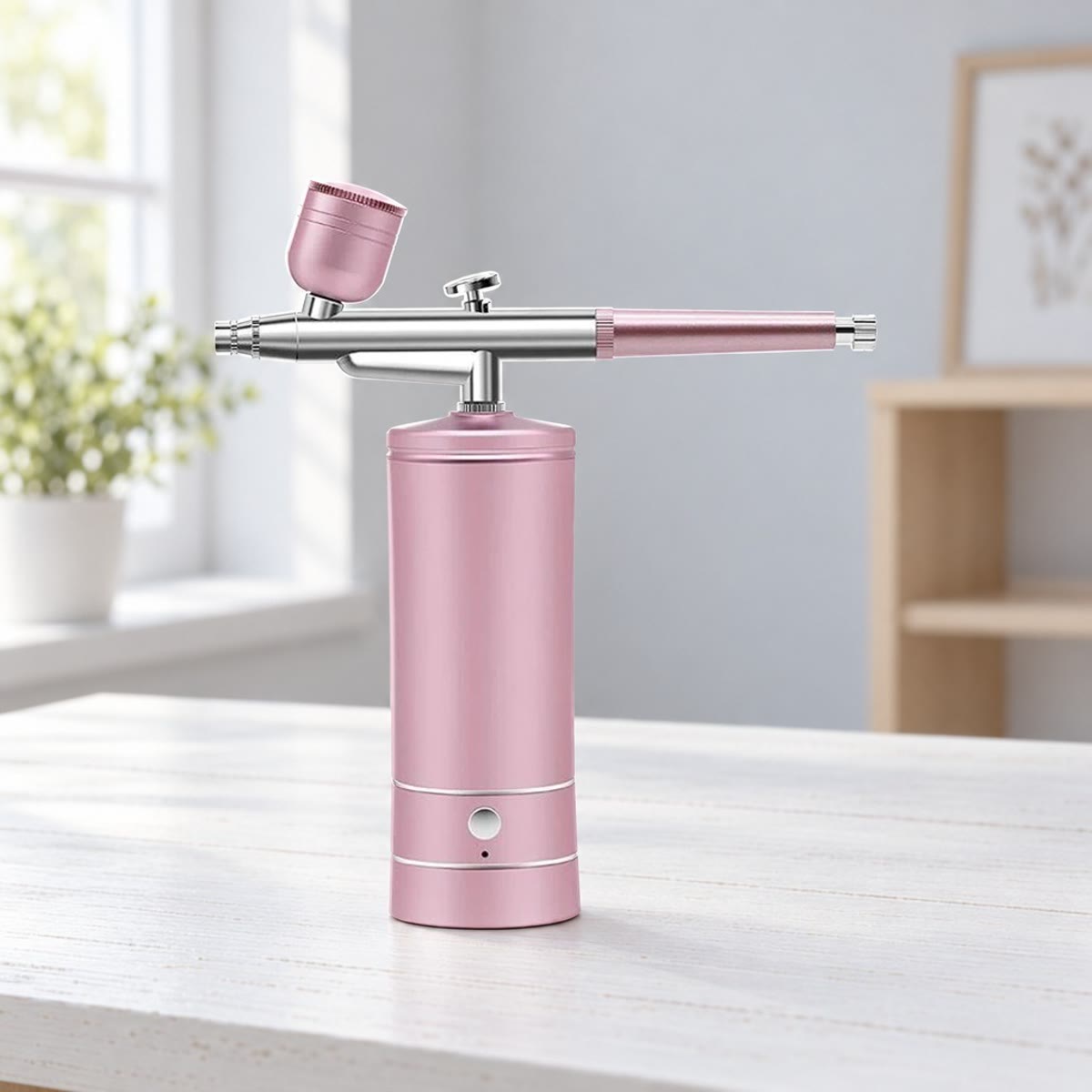 Rosa utgave av airbrushen med minikompressor på et hvitmalt skrivebord i et lyst rom