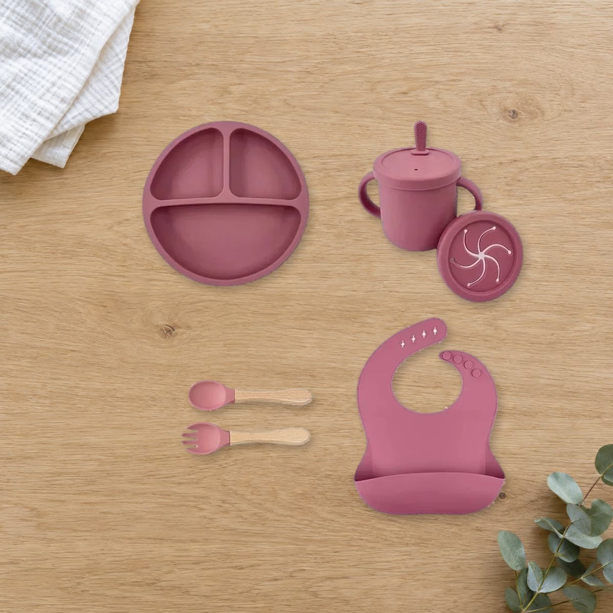 Barneservise i rosa silikon med tallerken, kopp, snacklokk, smekke, skje og gaffel lagt ut på et bord i eik