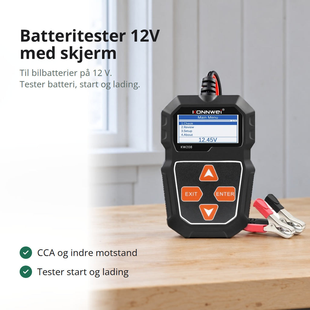 Svart batteritester 12V med skjerm og rød og svart klemme på en arbeidsbenk i en garasje, med snø utenfor vinduet