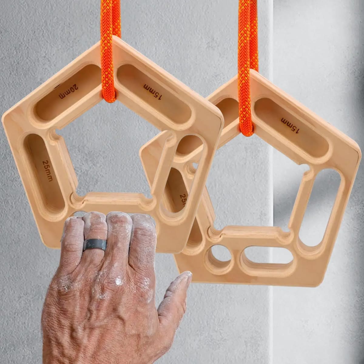 To femkantede hangboards i tau foran en grå vegg, med en hånd med klatrekritt på den nederste kanten