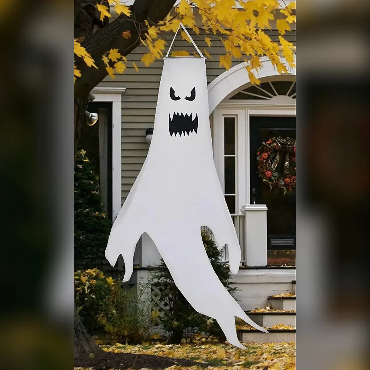 Hvitt halloween spøkelse med sint ansikt og takkete munn, hengende i et tre foran et hus på dagtid, uten lys