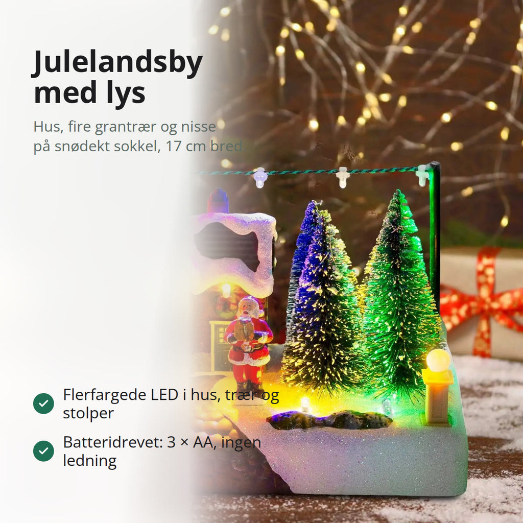 Julelandsby med lys: hus med snøtak, fire grantrær, nisse og lyslenke på en snødekt sokkel, tent på et bord