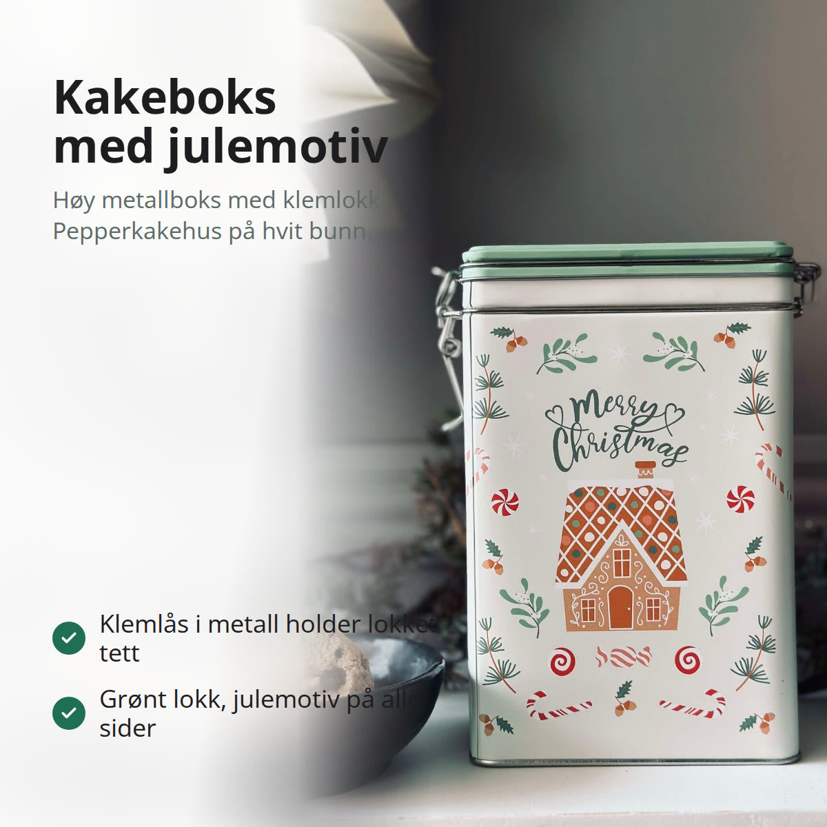 Høy kakeboks i metall med julemotiv, pepperkakehus og grønt klemlokk, ved siden av en skål med kjeks