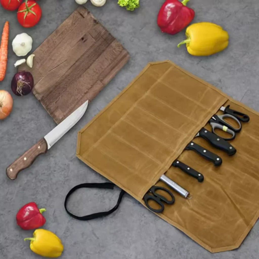 Knivmappe i kaki canvas åpen på en benk med kniver og saks i lommene, ved siden av grønnsaker og et skjærebrett