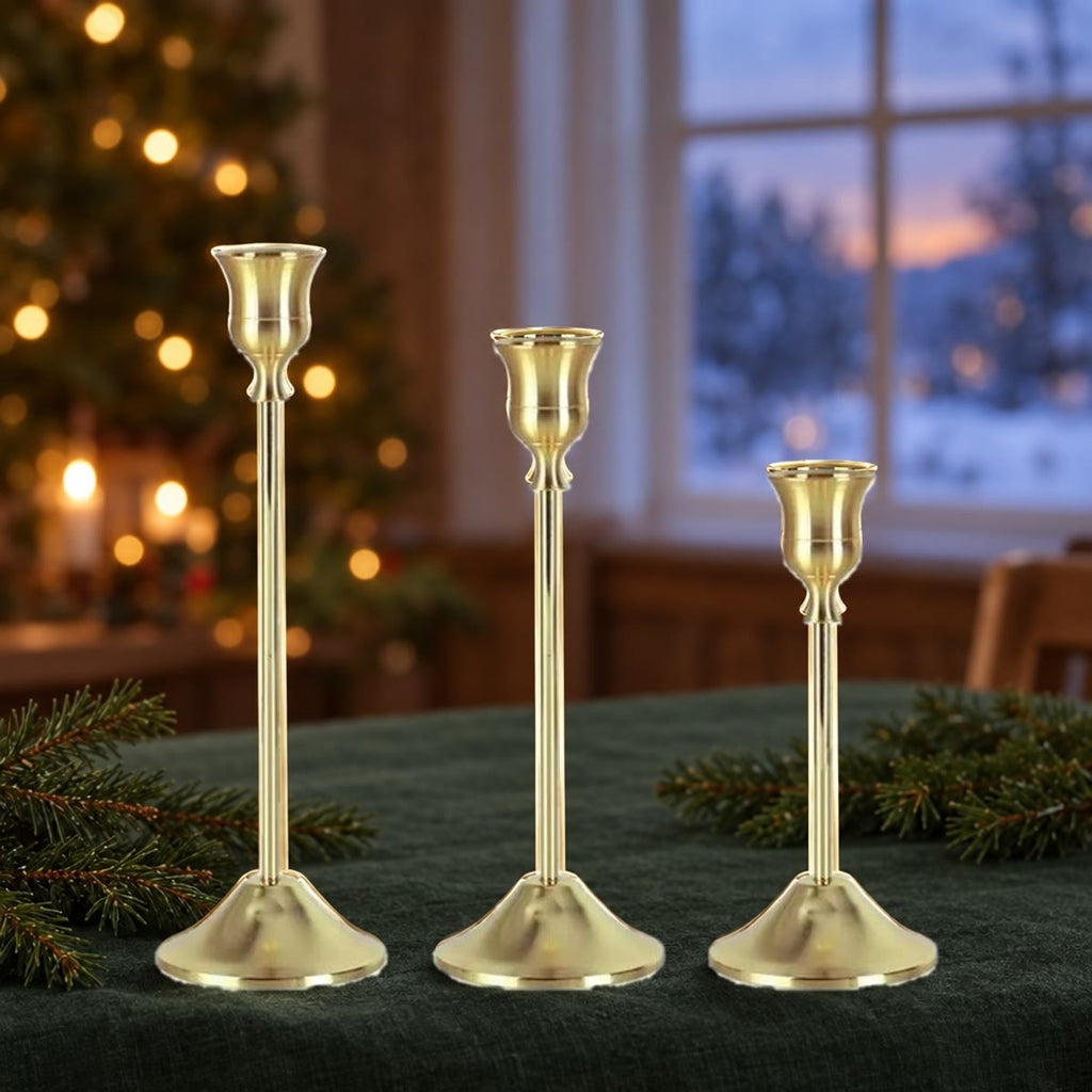 Tre lysestaker i gull, 22, 19 og 16 cm, på et julebord foran et pyntet juletre