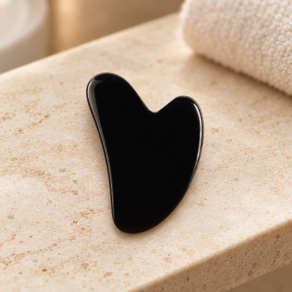Hjerteformet gua sha i sort obsidian som ligger på en travertinhylle på badet