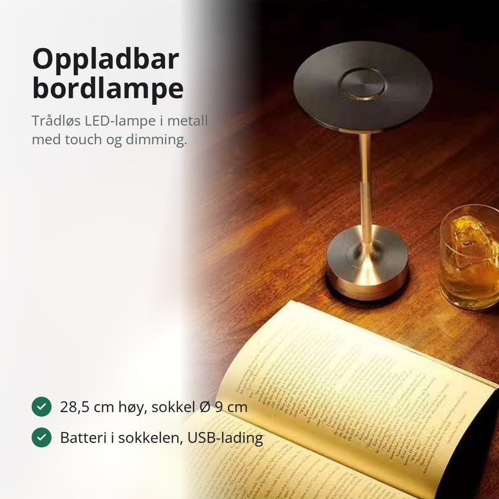 Oppladbar bordlampe i metall med flat, rund skjerm, tent på et trebord ved siden av en åpen bok og et glass