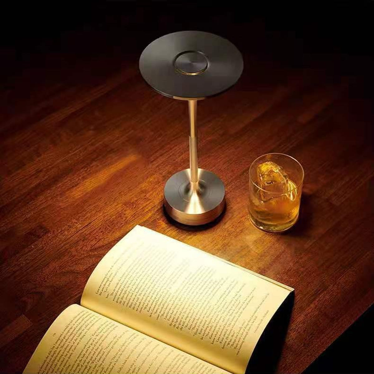 Oppladbar bordlampe tent på et mørkt trebord ved siden av en åpen bok og et glass