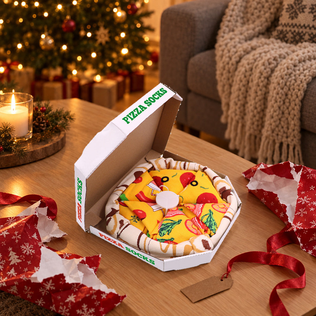 Åpen pizzaeske med fire par sokker rullet som pizzastykker på et salongbord ved juletreet, med revet gavepapir og rødt bånd