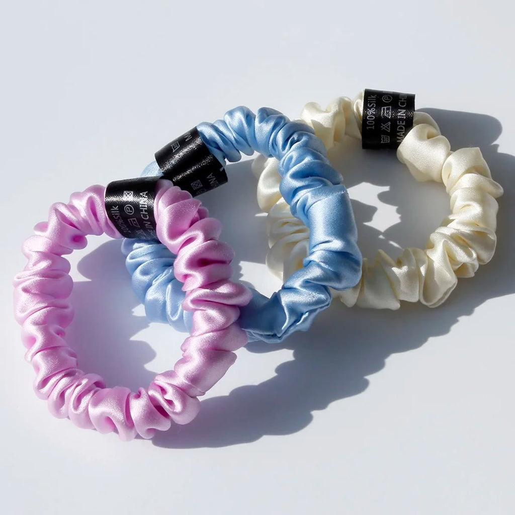 Tre silke scrunchies i rosa-lilla, lyseblått og krem med svart etikett, i sollys
