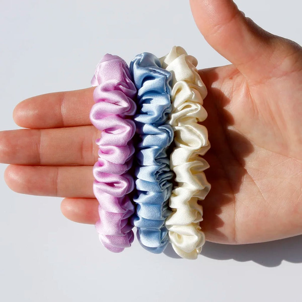 Hånd som holder tre smale silke scrunchies i rosa-lilla, lyseblått og krem