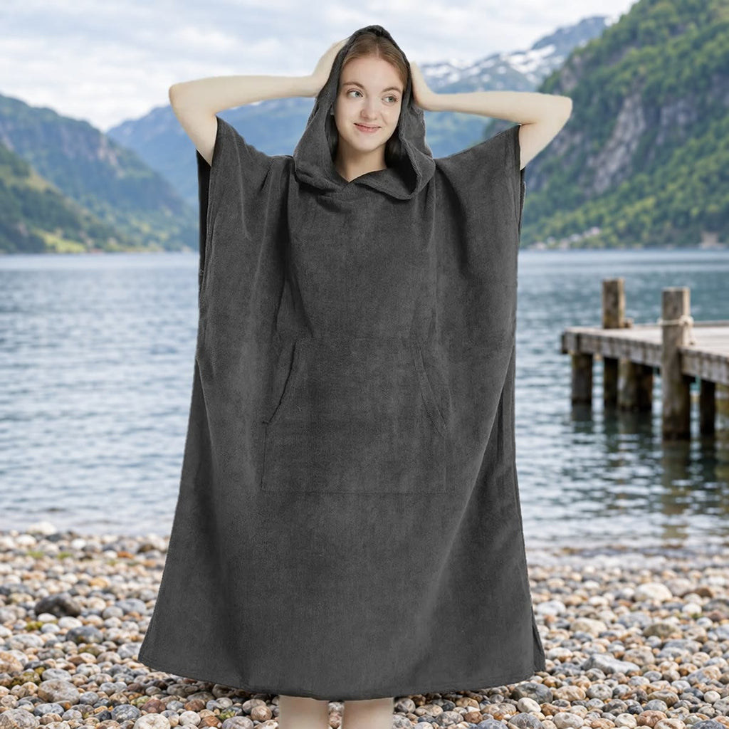 Kvinne i mørkegrå skifteponcho med hetten oppe på en steinstrand ved en norsk fjord, med brygge og fjell i bakgrunnen
