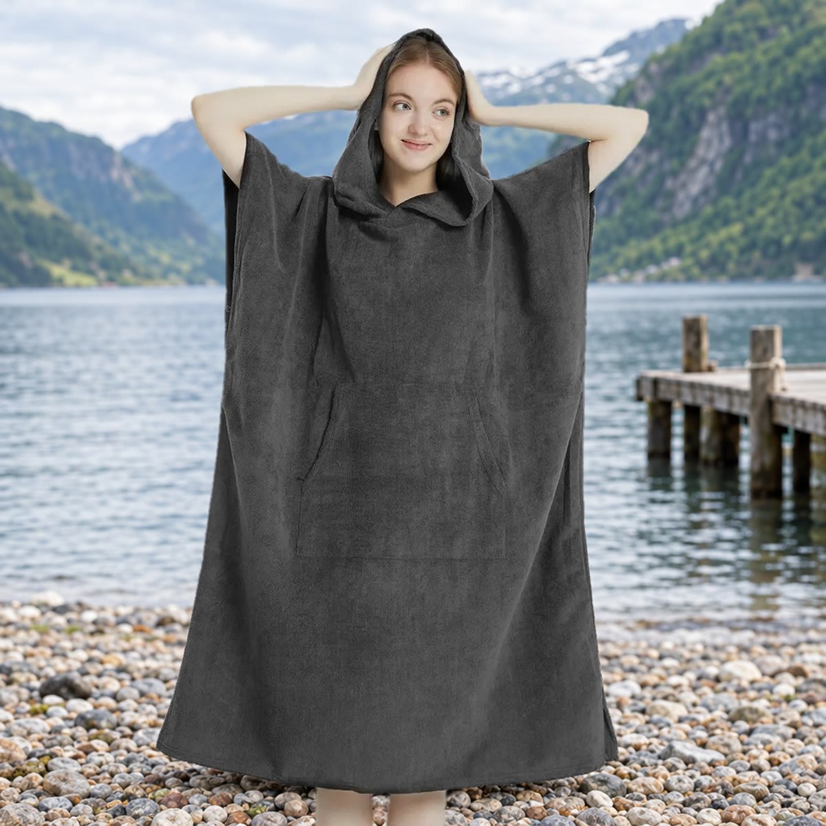 Kvinne i mørkegrå skifteponcho med hetten oppe på en steinstrand ved en norsk fjord, med brygge og fjell i bakgrunnen