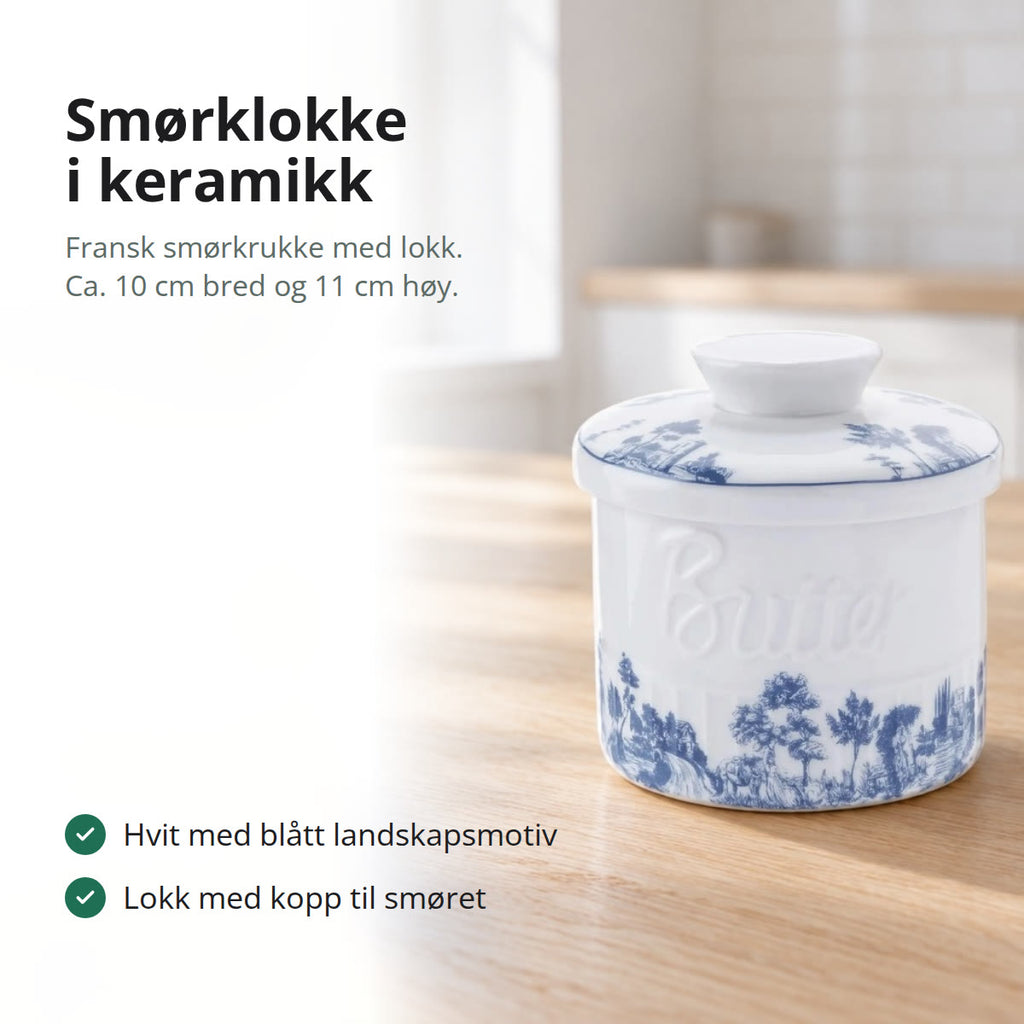 Hvit smørklokke i keramikk med blått landskapsmotiv og ordet Butter i relieff, på et kjøkkenbord i lys eik