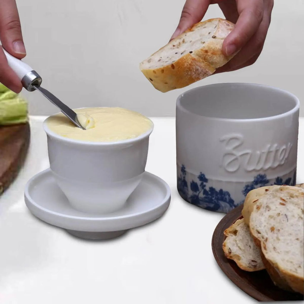 En hånd tar smør med kniv fra koppen på det omsnudde lokket; krukken med blått motiv og ordet Butter står ved siden av