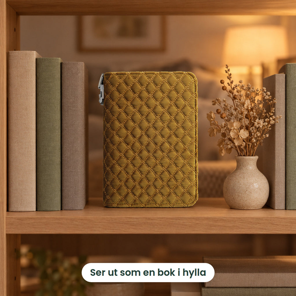 Smykkeboken i gull stående i en bokhylle mellom bøker og en vase med tørkede blomster