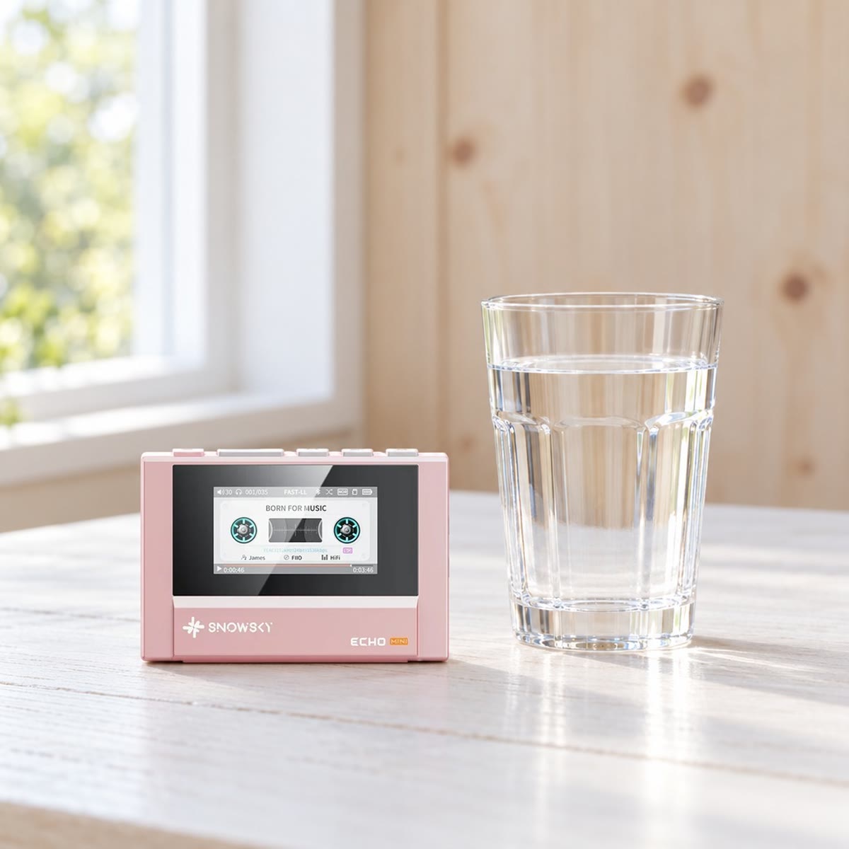 Rosa SnowSky Echo Mini stående på et lyst bord ved et vindu, ved siden av et vannglass, med kassettspoler på skjermen