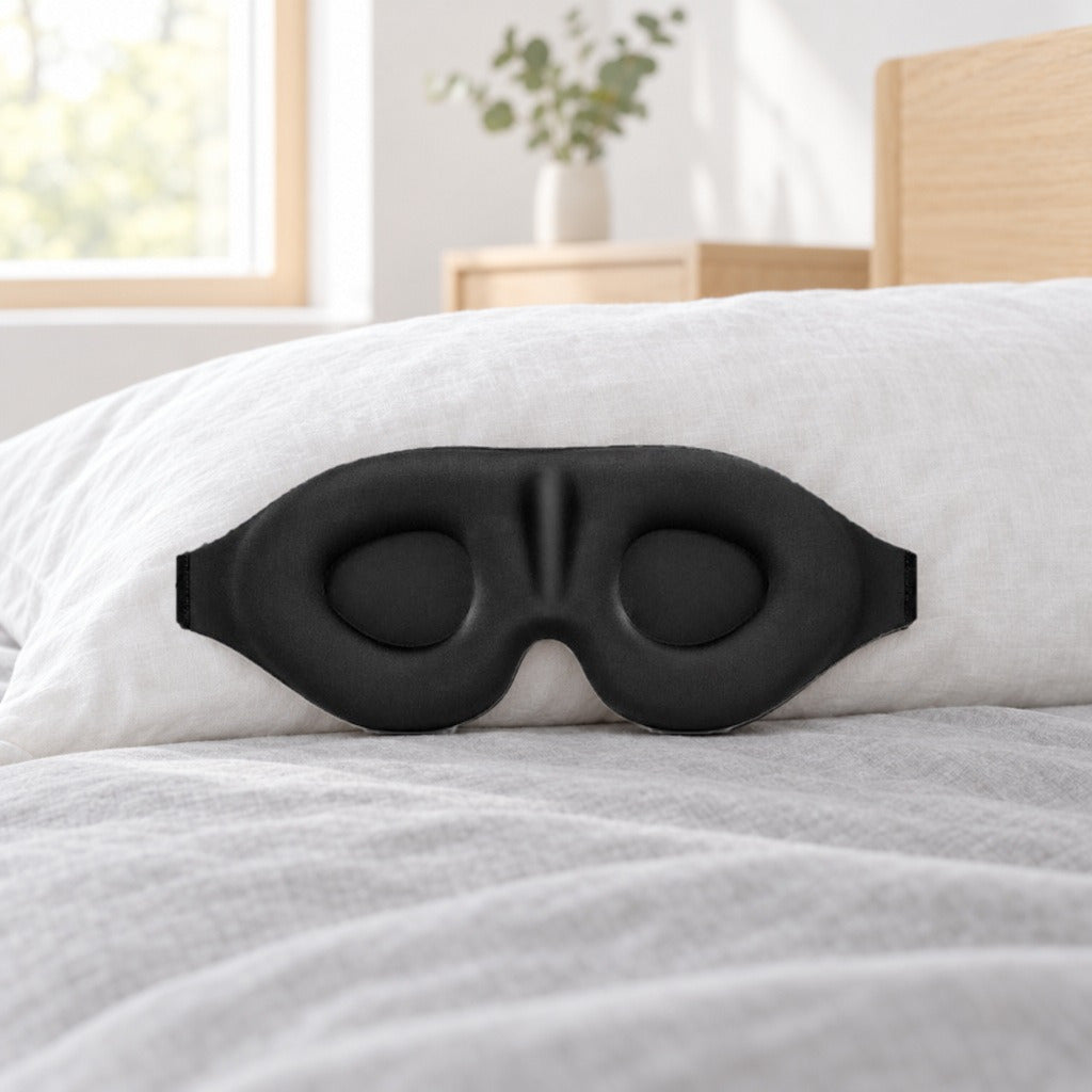 Svart 3D-sovemaske med ovale øyekopper og formet nesebro, lent mot en hvit pute på en seng