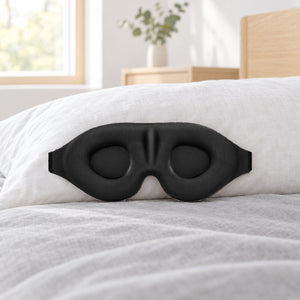 Svart 3D-sovemaske med ovale øyekopper og formet nesebro, lent mot en hvit pute på en seng