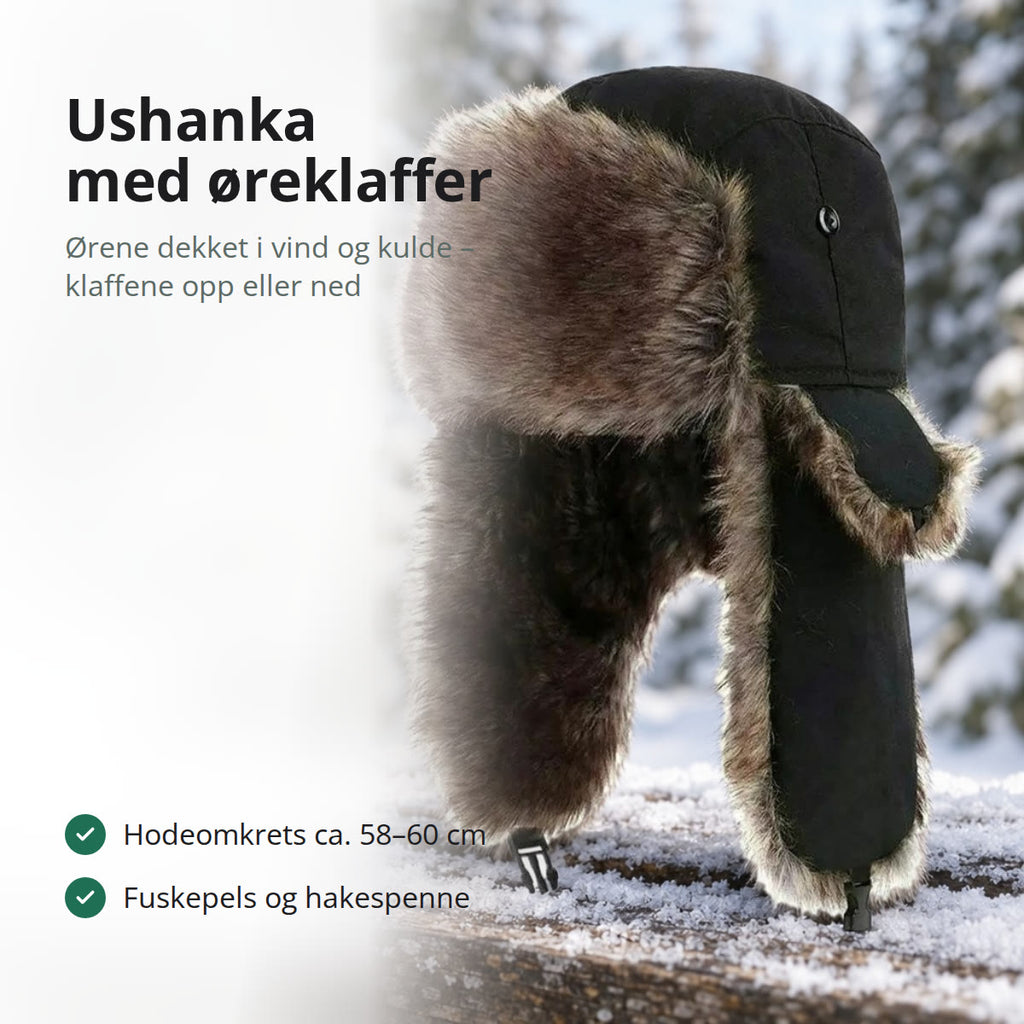 Svart ushanka med øreklaffer i gråbrun fuskepels på et snødekt rekkverk av tre foran granskog
