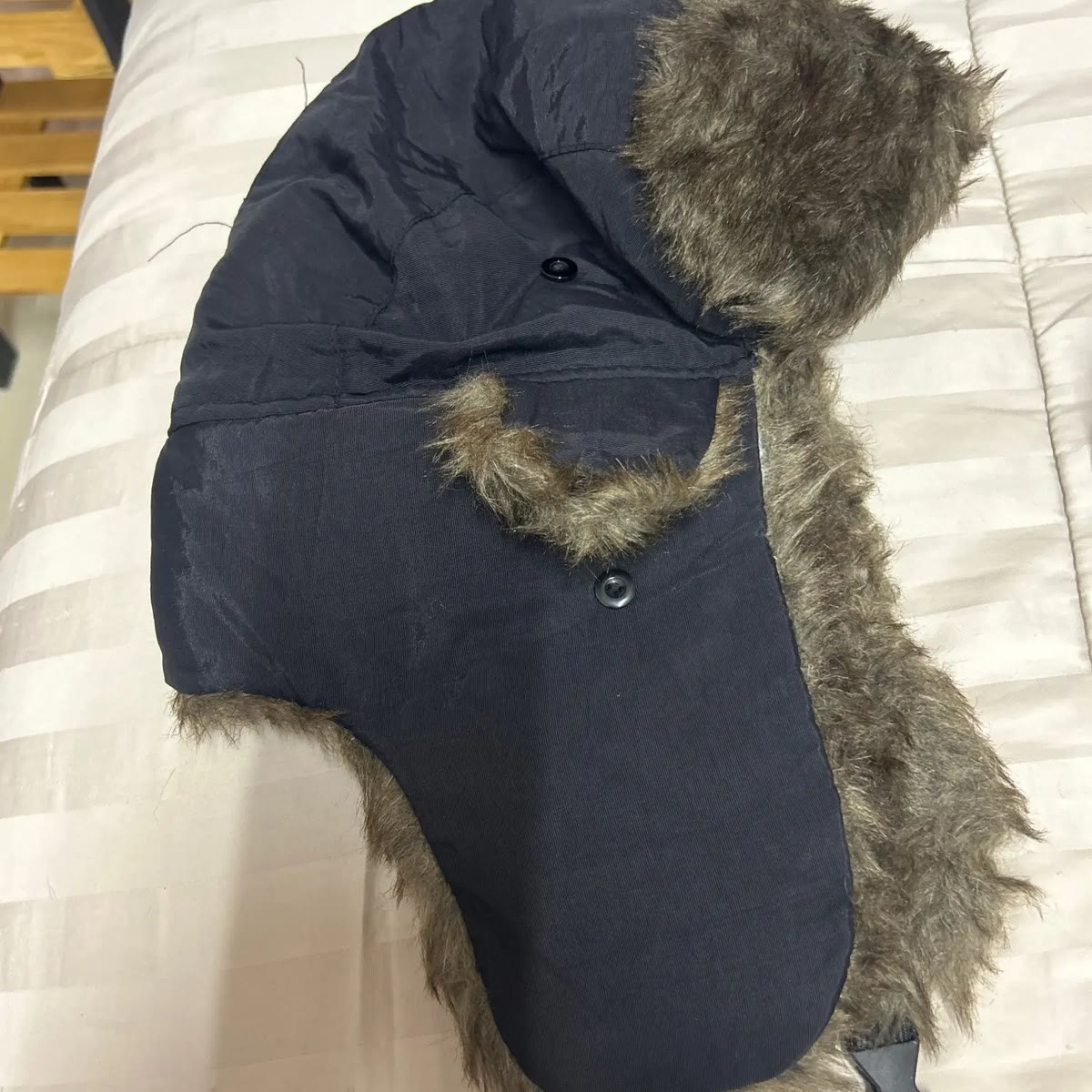 Kjøperbilde av ushankaen lagt flatt på en seng, med svart ytterstoff, fuskepels og spennen nederst på klaffen