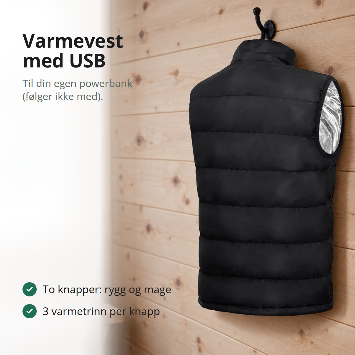Svart varmevest sett bakfra, hengende på en knagg på en lys panelvegg i en hytte, med snødekte grantrær utenfor vinduet