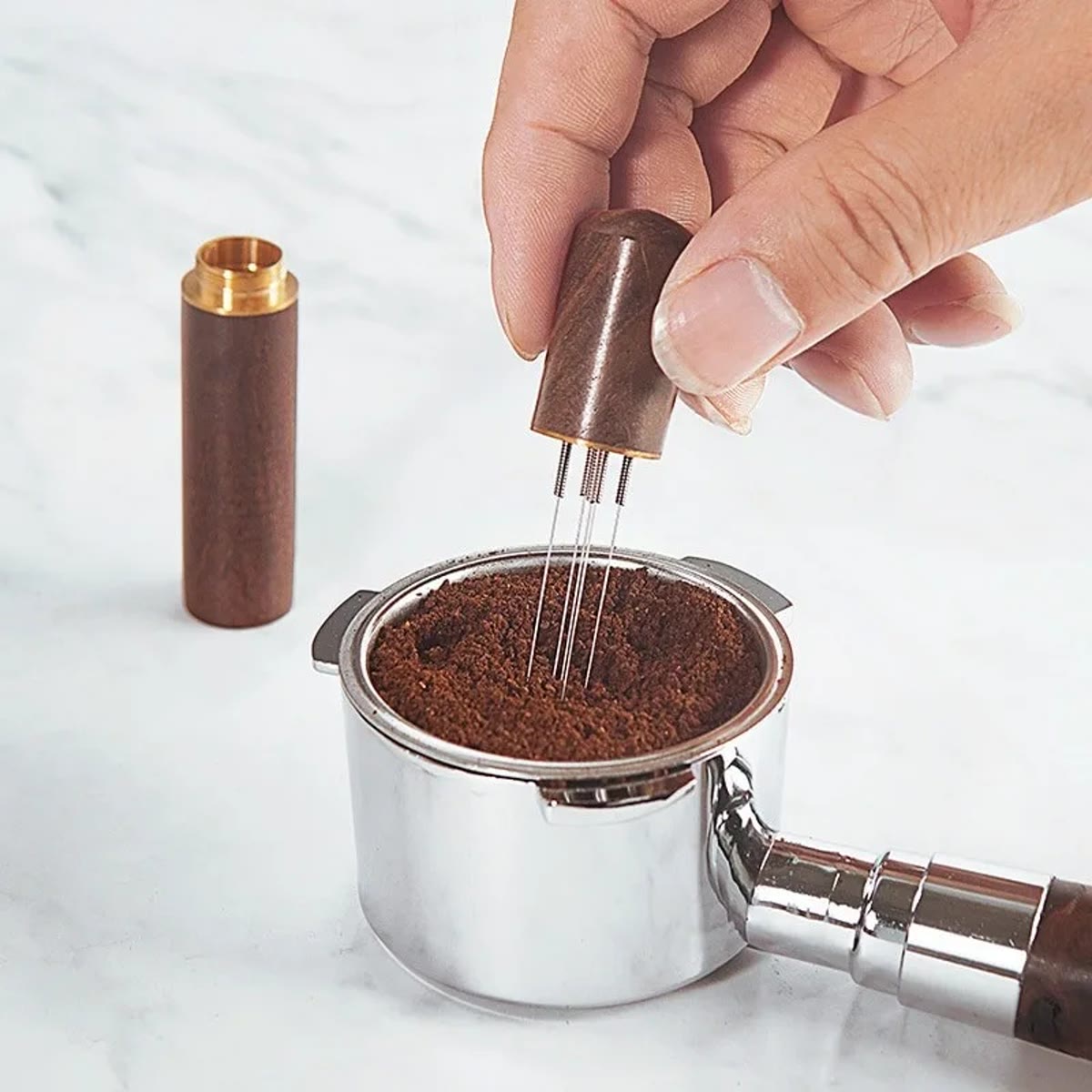 Hånd som rører med WDT-verktøyet i kaffepulveret i en filterkurv, med lokket ved siden av