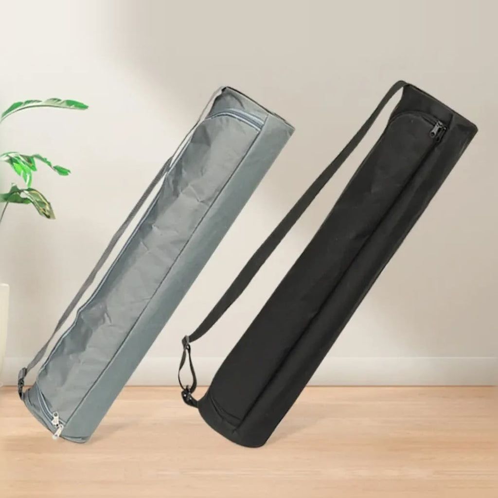 En grå og en svart yogamatte bag med skulderstropp, lent mot en lys vegg på et tregulv